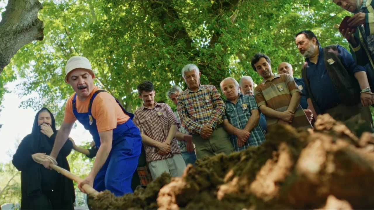 Paranormal Cuma (2024) movie still: medium — A group of people stands among trees at a burial site during the day. A man in work clothes uses a…