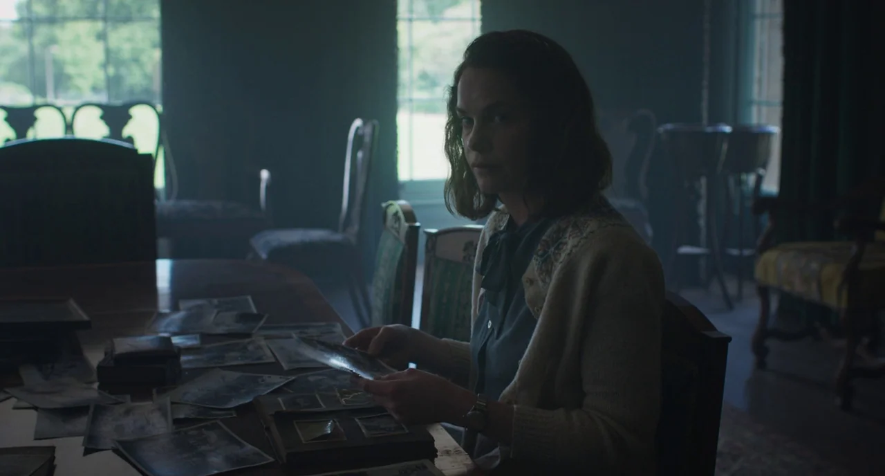 The Little Stranger (2018) movie still: medium — A dimly lit room contains a long dark wooden table covered with vintage photographs. A woman sits…