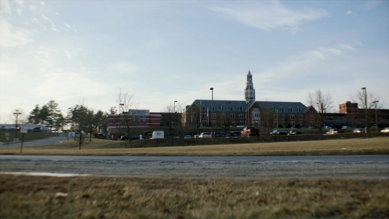 What Happened at Fells Acres? (2023) movie still: establishing — A large brick school complex sits on a grassy hill under a clear sky. A paved road…