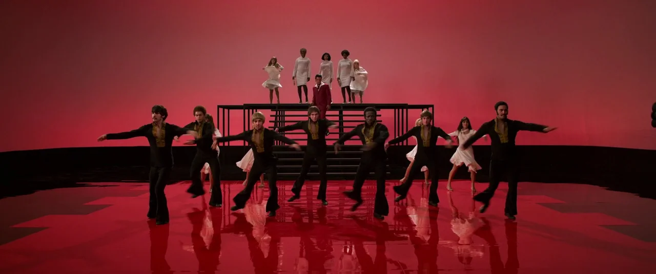 Elvis (2022) movie still: wide — A minimalist studio stage with a glossy red floor and deep red background wall. A group of dancers performs…