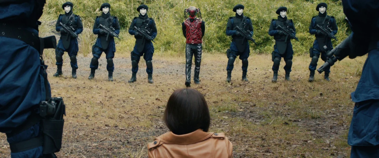 Shin Kamen Rider (2023) movie still: wide — A field of tall dry grass surrounds a group of soldiers in blue tactical gear. A line of soldiers stands…