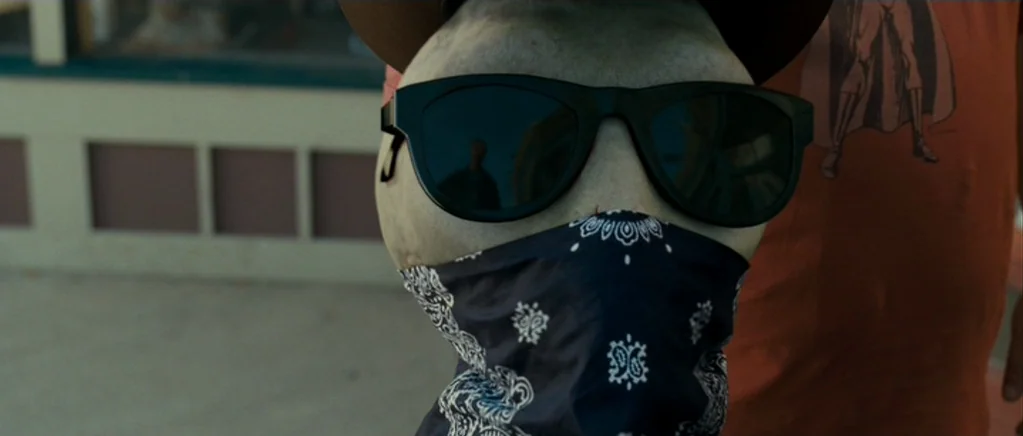 Paul (2011) movie still: closeup — A close-up view shows a masked figure wearing dark sunglasses on a public walkway. The subject's face is covered…