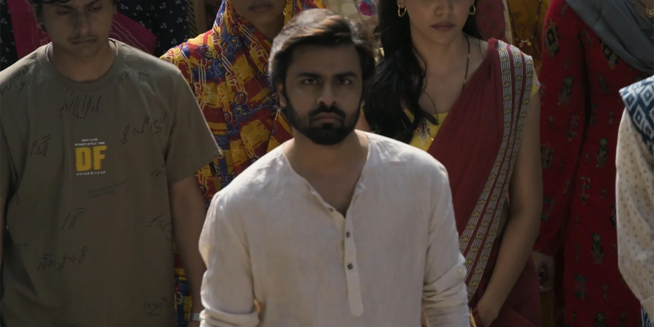 Dry Day (2023) movie still: medium — A group of people stands in a daylight outdoor setting with colorful textiles visible. A man in a beige shirt…