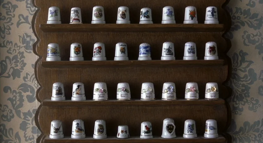 About Schmidt movie still: closeup — A wooden wall-mounted rack displays rows of ceramic thimbles with various painted designs. The display is…