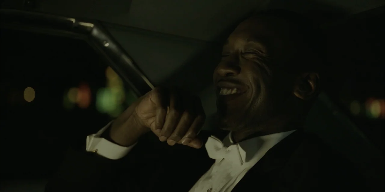 Green Book (2018) movie still: closeup — The interior of a car at night shows a man dressed formally for an event. He is laughing with his head…