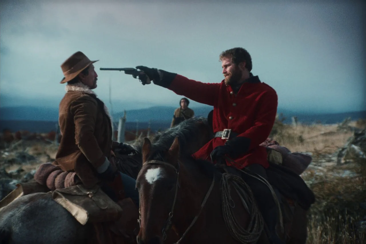 The Settlers (2023) movie still: medium — Two men on horseback face each other in an open, desolate, and hilly landscape. One man in a red coat…