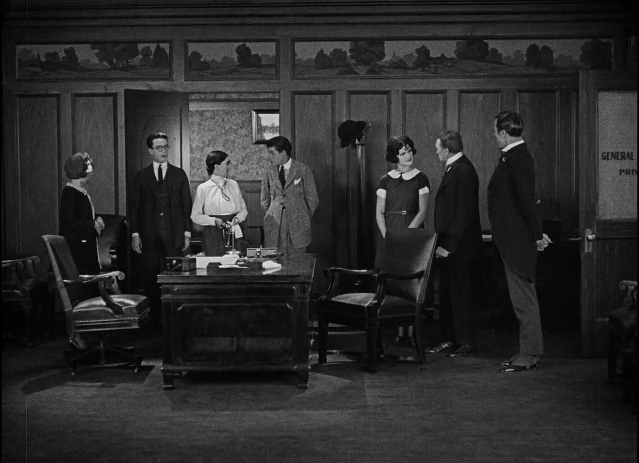 Safety Last! (1923) movie still: fullbody — A wood-paneled office from the 1920s features a large central desk and wall art. Six people stand in the…