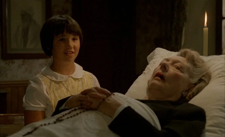 Milou en Mai (1990) movie still: medium — A dim bedroom contains an elderly woman resting in a bed with a white pillow. A young girl sits beside the…