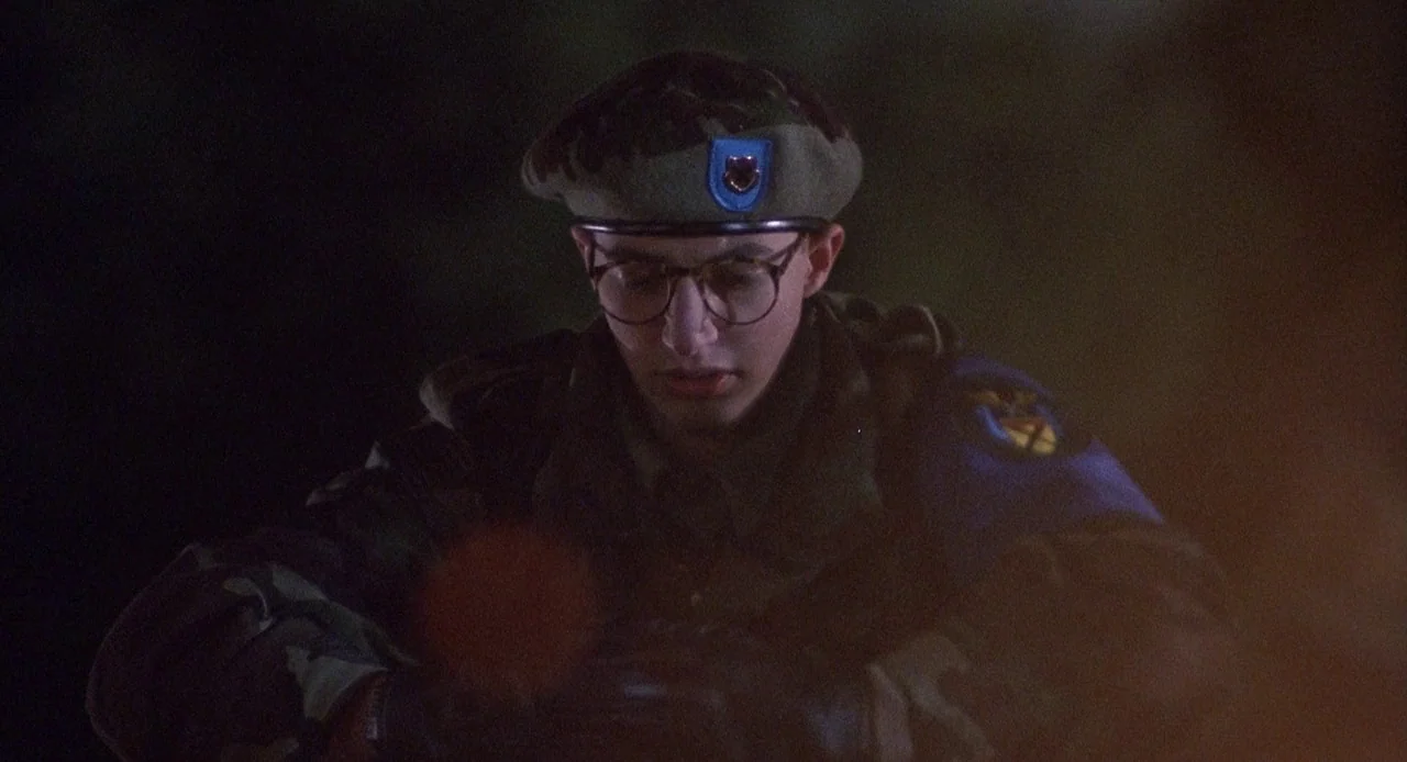 Child’s Play 3 (1991) movie still: medium — A dark outdoor setting at night with a soldier wearing camouflage in the foreground. The soldier wears…