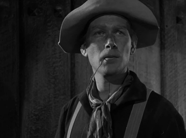 Rio Grande (1950) movie still: closeup — The interior of a wooden barn shows vertical planks of aged timber in darkness. A man wearing a…