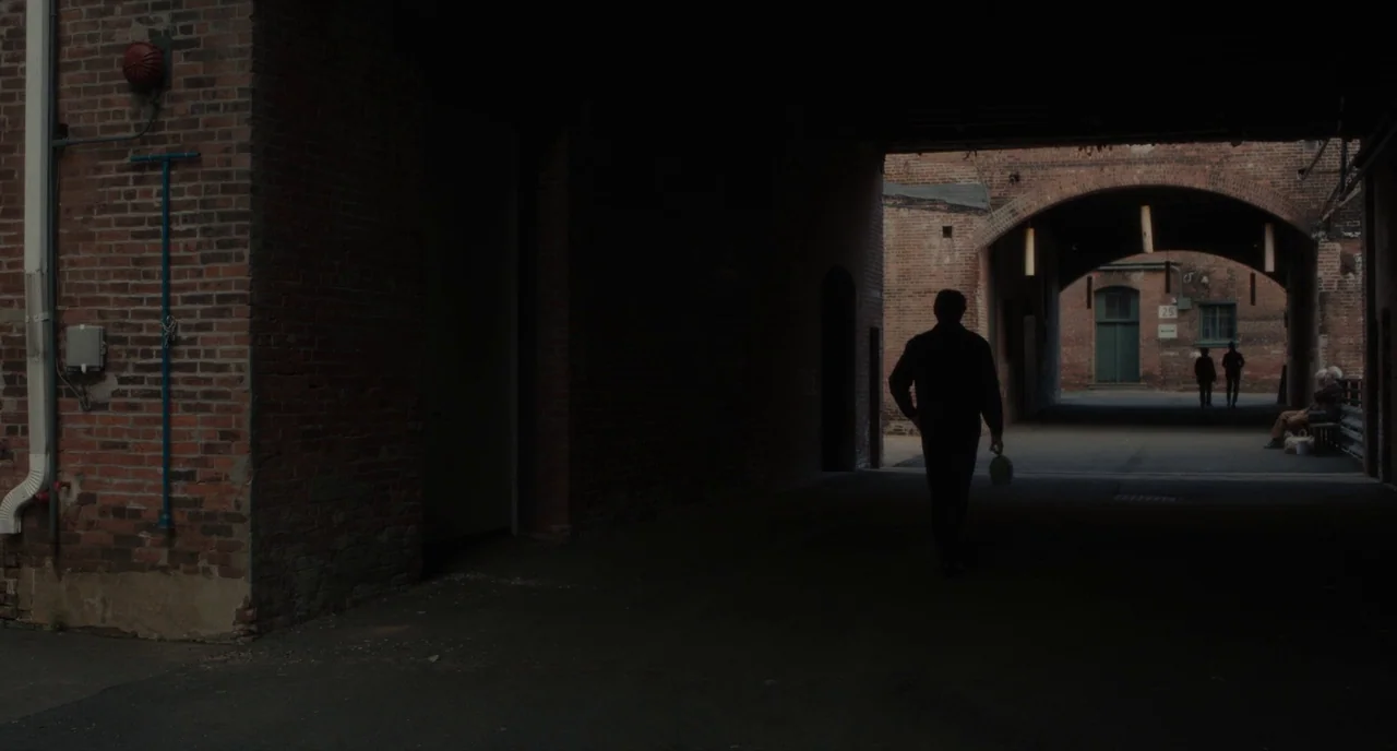 Paterson (2016) movie still: wide — A dark brick tunnel serves as a passage between two areas of industrial brick buildings. A man walks away from…