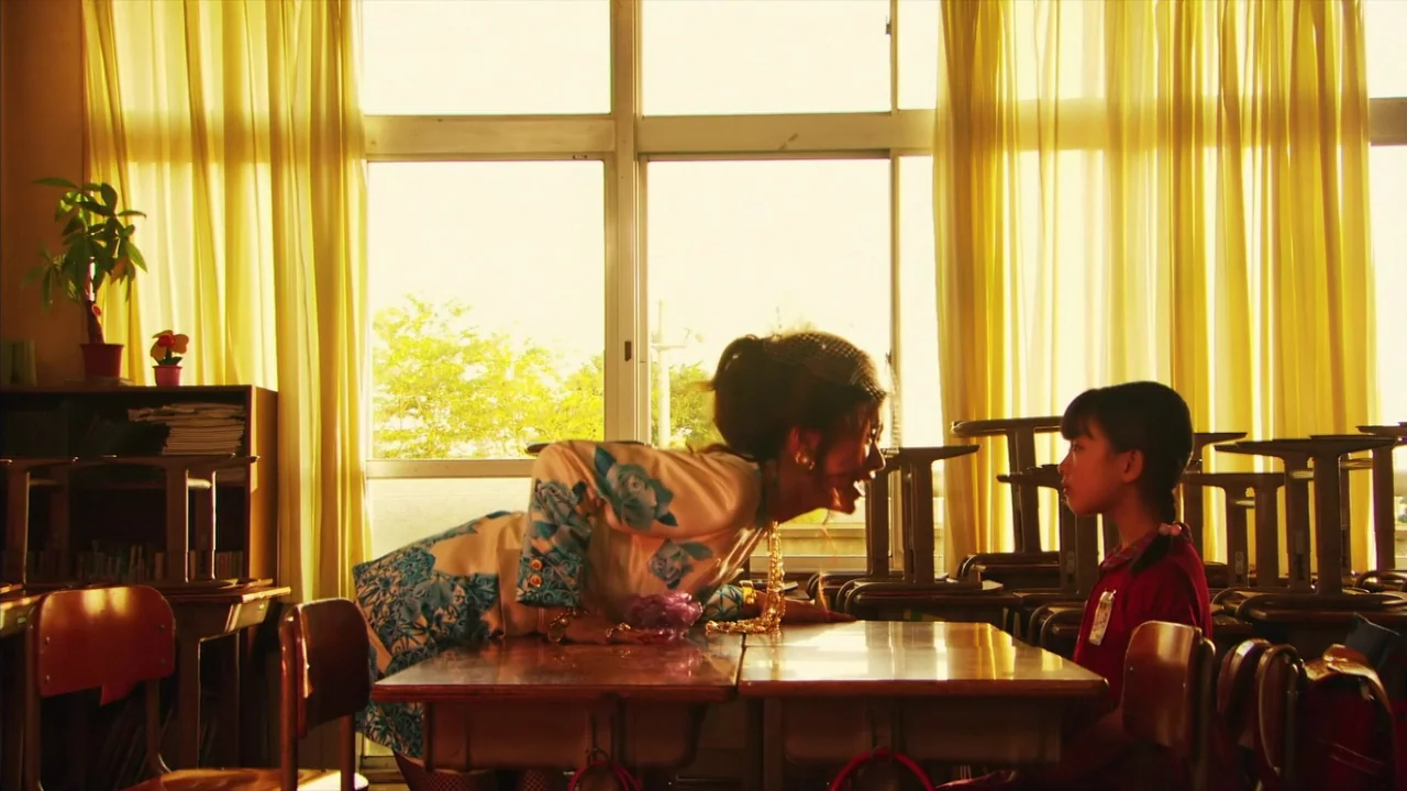 Kamikaze Girls (2004) movie still: medium — A sunny, yellow-toned classroom with empty wooden desks and bright drawn curtains. An adult woman leans…