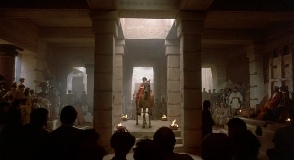 Time Bandits (1981) movie still: wide — A stone hall with square pillars hosts a crowd watching a rider on horseback. Sunlight pierces through an…