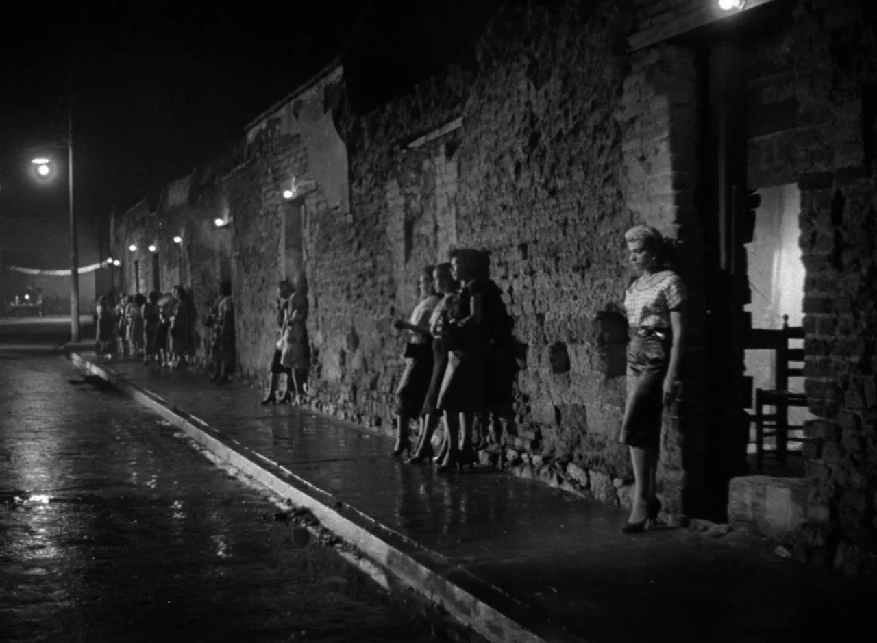 Victims of Sin (1951) movie still: wide — A wet concrete sidewalk runs alongside a long, crumbling exposed brick wall at night. Several women are…