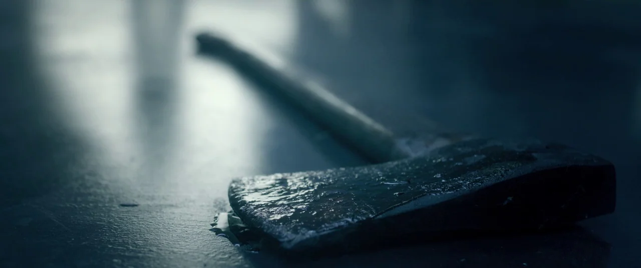 The Void (2016) movie still: closeup — A metal axe head lies on a dark, wet reflective floor surface. Liquid droplets surround the blade head in the…
