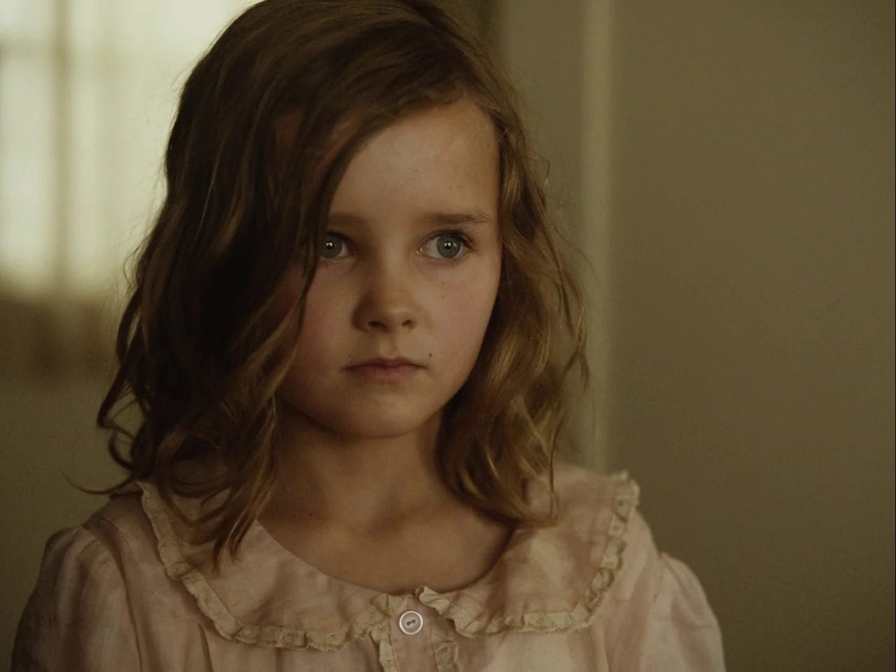 Blonde (2022) movie still: closeup — A dimly lit room with plain walls provides a neutral backdrop for the portrait. A young girl in a pale pink…