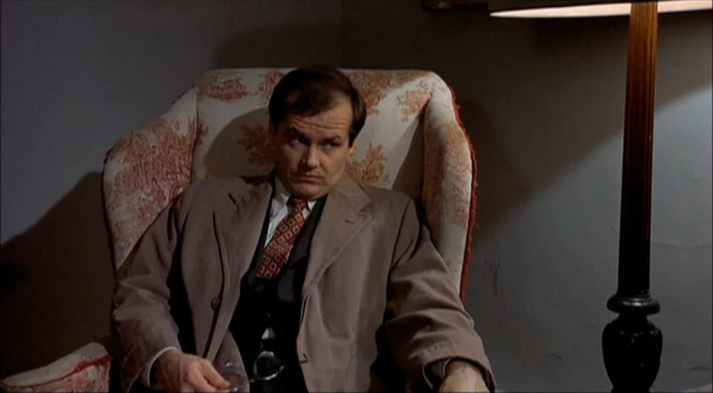 The King Of Marvin Gardens movie still: medium — A dimly lit room features a man sitting in a patterned fabric armchair. The man holds his glasses…