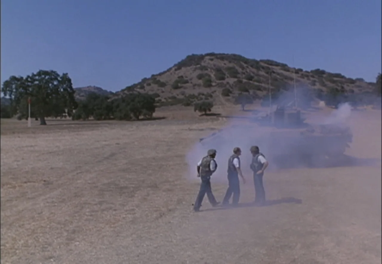 The Pentagon Wars (1998) movie still: wide — A dry open field features rolling hills and a smoking military vehicle. Three men wearing tactical gear…