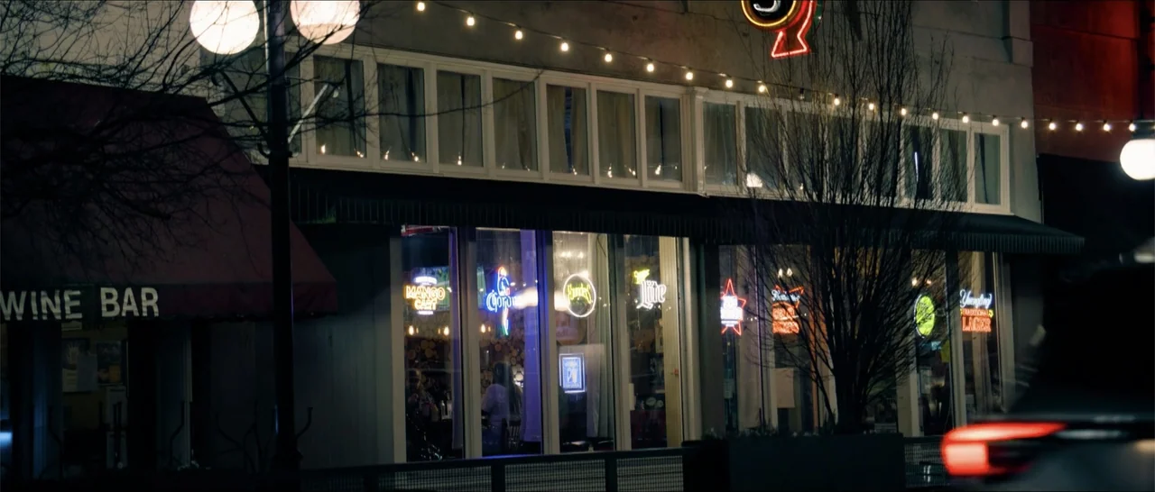 Crescent City (2024) movie still: establishing — A commercial building front features large glass windows adorned with multiple glowing neon beer…