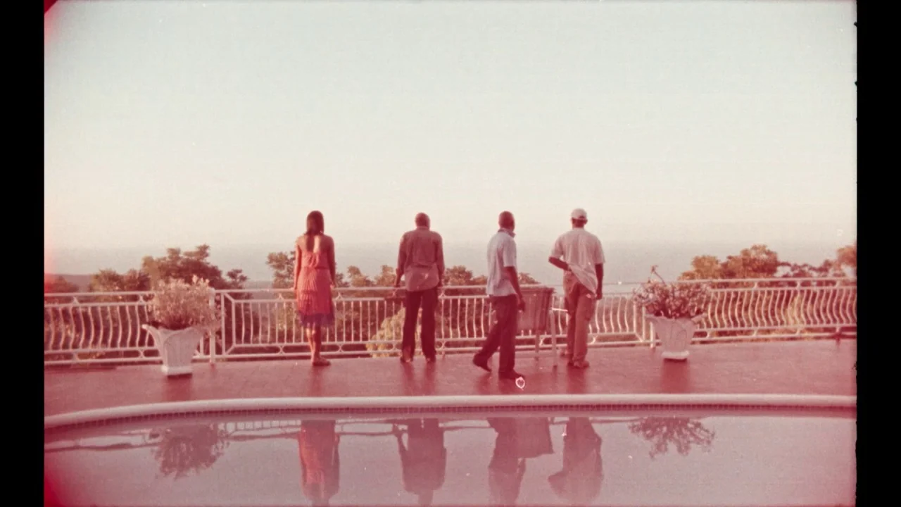 Black Mother (2018) movie still: wide — A spacious outdoor terrace with a white railing overlooks a distant horizon during daytime. Four people…
