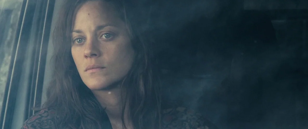 Rust And Bone (2012) movie still: closeup — A woman sits inside a car while looking out through a window. The glass shows reflections of the…