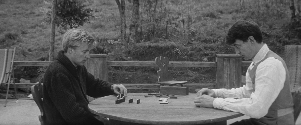 Jules and Jim (1962) movie still: medium — A rustic wooden patio overlooks a grassy outdoor area with scattered trees. Two men sit across from each…