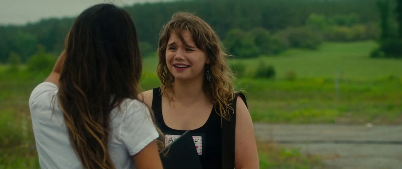 Random Acts of Violence (2019) movie still: medium — A rural landscape with a paved road and lush green foliage in the background. Two young women…