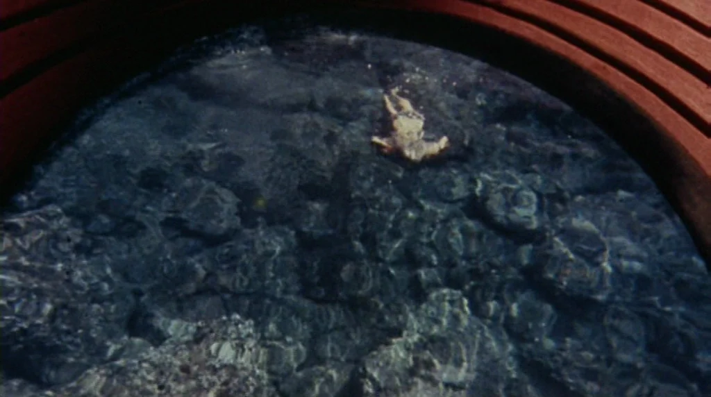 Danger Diabolik (1968) movie still: wide — A dark rocky pool is viewed through a circular wooden frame. A small figure floats motionless on the…