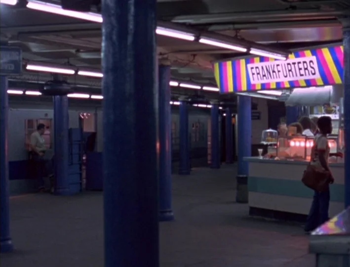 News From Home (1977) movie still: wide — A subway platform features blue pillars and a snack stand labeled with colorful signage. A man stands at…
