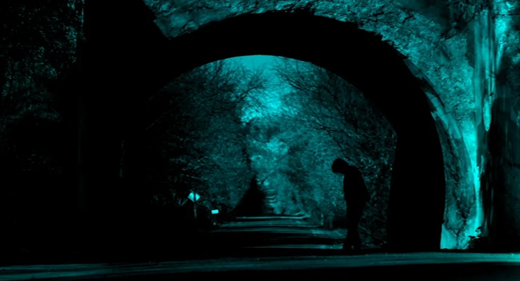 Mama (2013) movie still: wide — A dark stone tunnel is illuminated by artificial cyan light reflecting off its surfaces. A single person stands in…