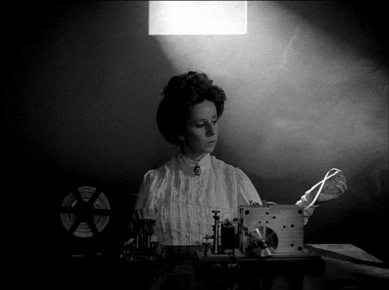 My 20th Century (1989) movie still: medium — A woman works at a wooden desk with historical telegraph equipment against a dark background. She…