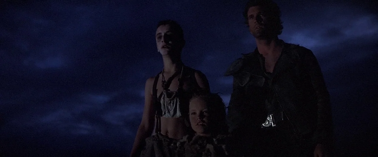 Mad Max Beyond Thunderdome (1985) movie still: medium — Three individuals stand in a dark, open area at night under a blue sky. The man, a teenager,…