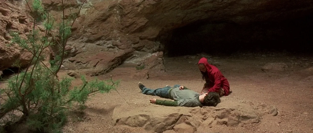 The Hills Have Eyes (2006) movie still: wide — A dry desert cave entrance with red rock formations and sparse green brush. A woman in a red hoodie…