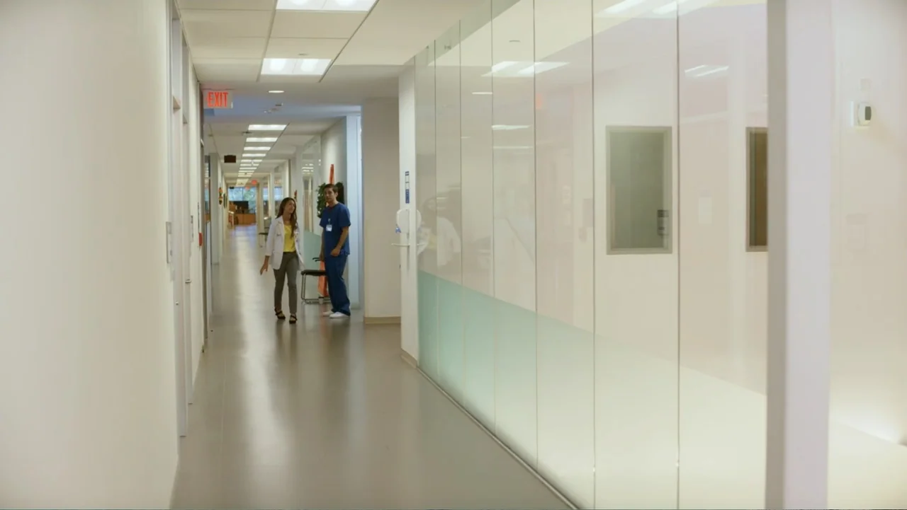 Christmas with a Prince (2018) movie still: fullbody — A modern hospital hallway with glossy floors and a long glass partition wall. A man and a…