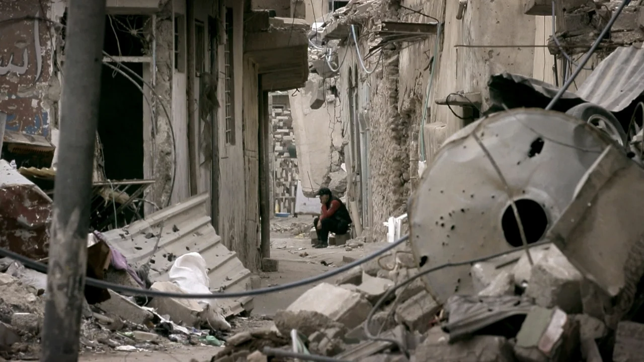 City of Two Springs (2019) movie still: wide — A narrow, rubble-strewn alleyway is flanked by damaged, deteriorating concrete buildings. A lone…