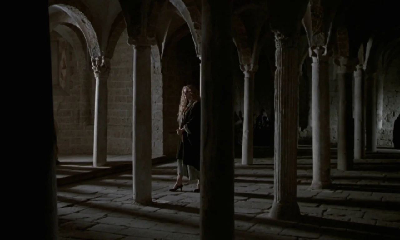 Nostalghia (1983) movie still: fullbody — A stone cloister features long rows of rounded arches supported by thick masonry columns. A woman walks…