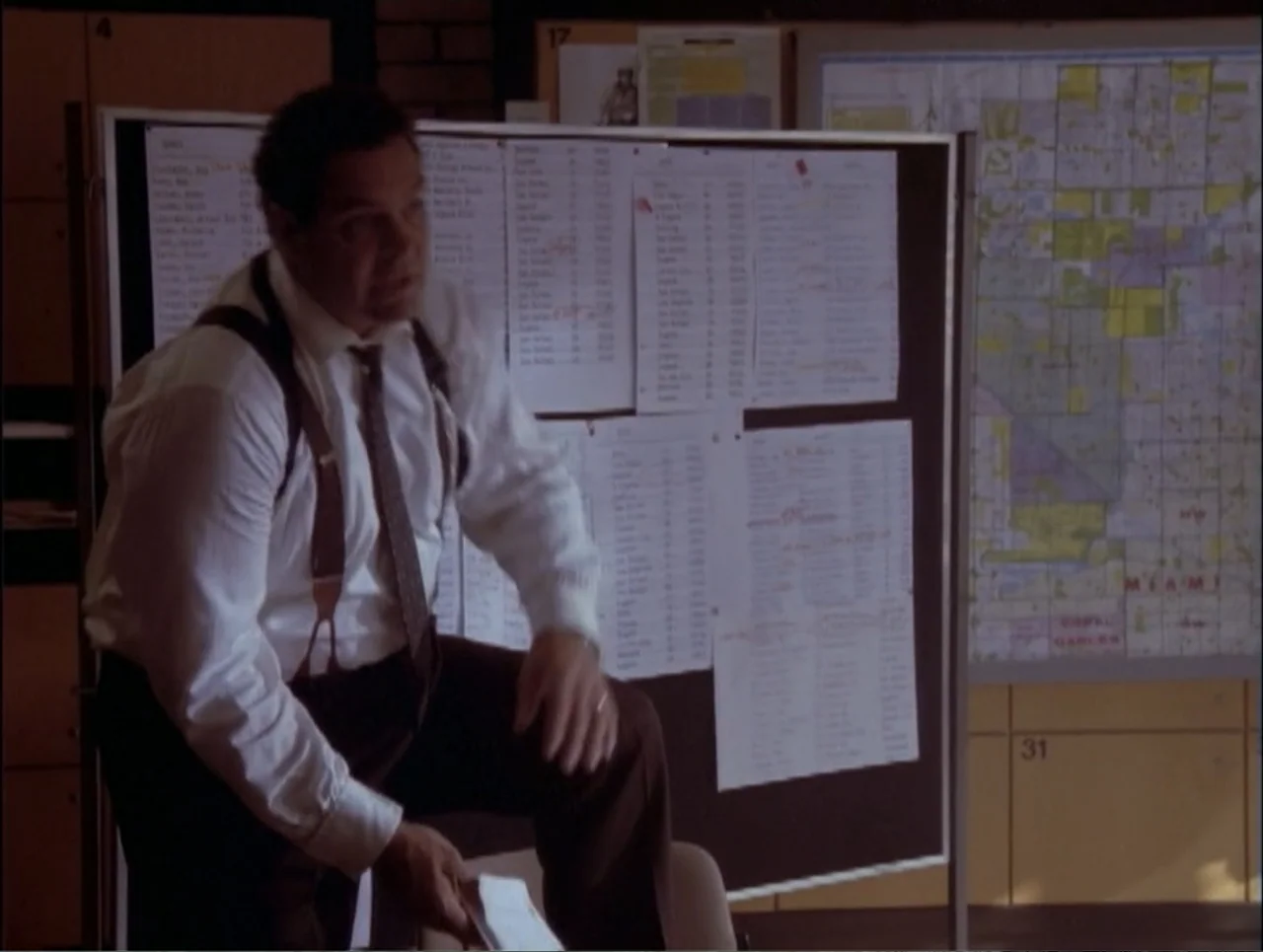 Code Name: Wolverine (1996) movie still: medium — A workspace features a large black bulletin board covered in paper lists and maps. A man in a…