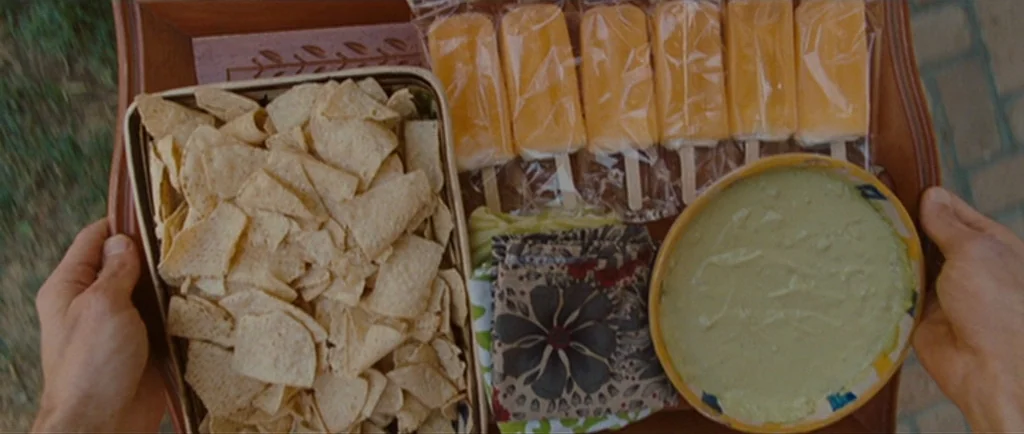 Greenberg (2010) movie still: closeup — A wooden tray holds a basket of tortilla chips, orange popsicles, and green dip. A person's hands are…
