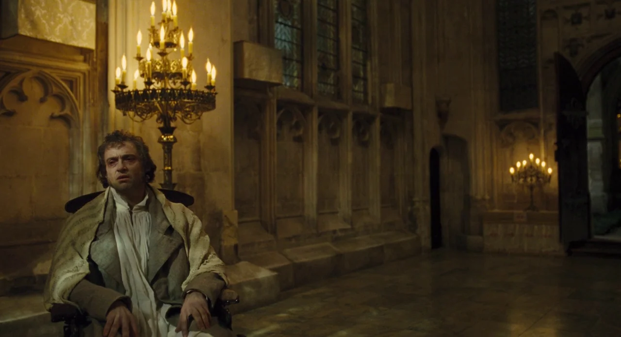 Les Misérables (2012) movie still: medium — An ornate stone room with high vaulted arches and stained glass windows. A man in period clothing sits…