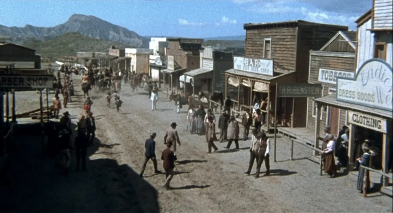 Chato's Land (1972) movie still: wide — A dusty 19th-century frontier town street is lined with wooden buildings featuring hand-painted signs.…