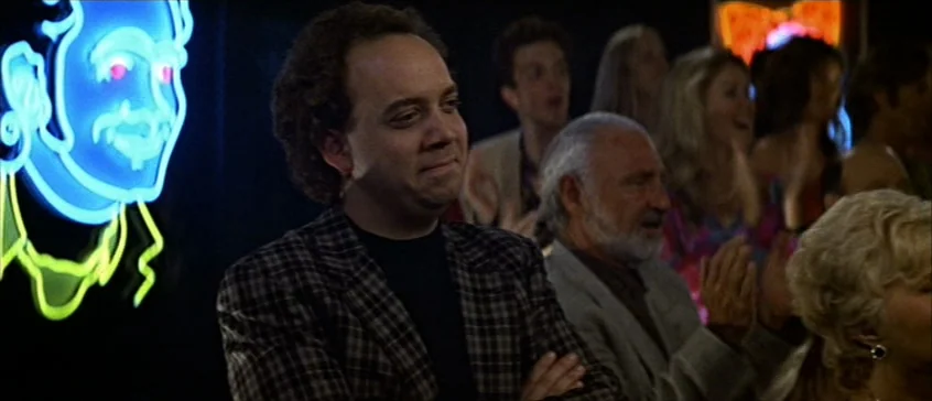 Man On The Moon (1999) movie still: medium — A dimly lit club environment features a prominent blue neon face sign on the wall. A man in a checkered…