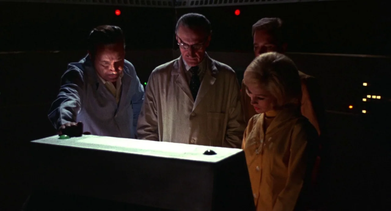 Queen of Blood (1966) movie still: medium — A dimly lit technical workspace is dominated by a central rectangular console panel. Four people in…