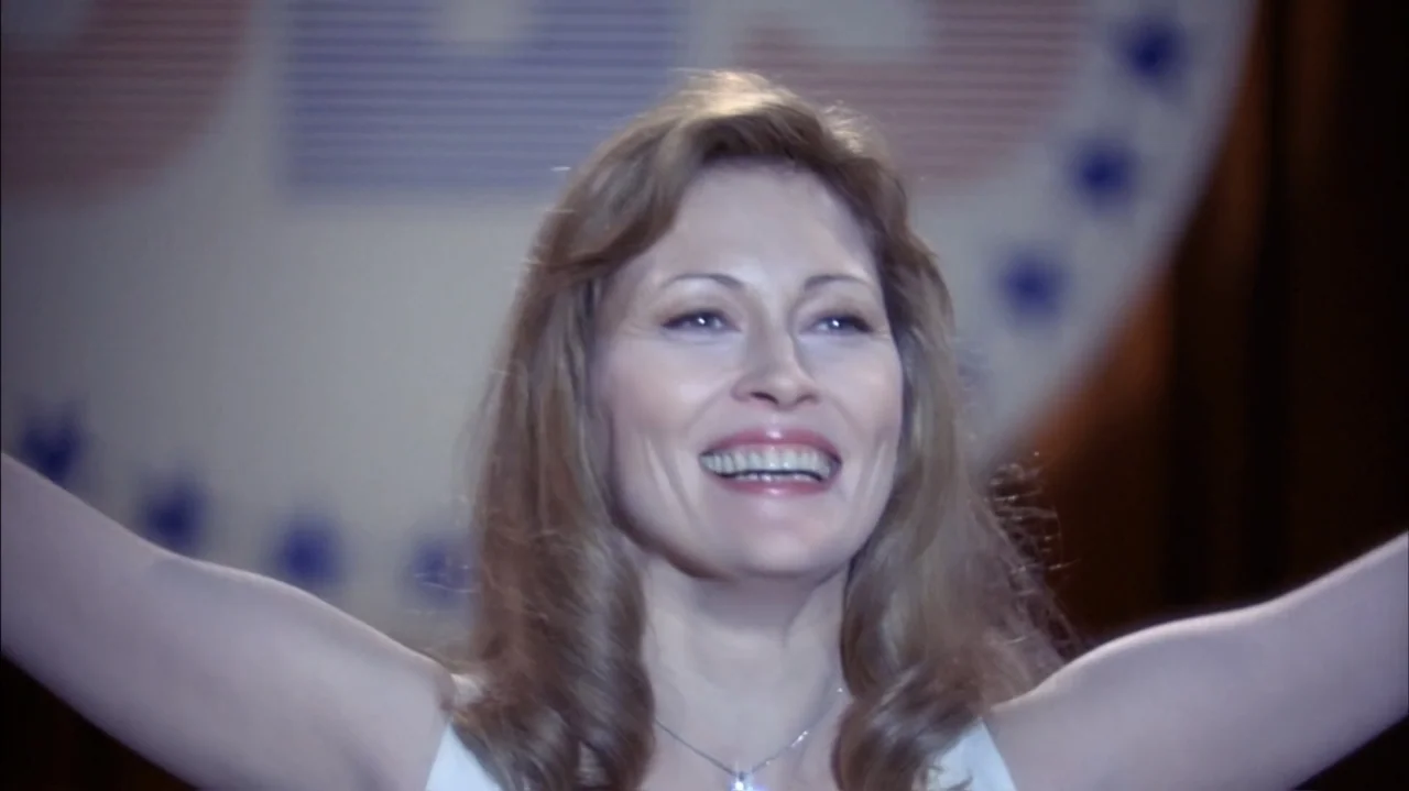 Faye (2024) movie still: closeup — A woman stands on a bright indoor stage with a blurred political backdrop. She smiles broadly while raising both…
