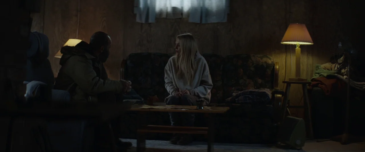Hold The Dark (2018) movie still: medium — A dimly lit room with plywood walls, floral patterned furniture, and two warm table lamps. A man and a…