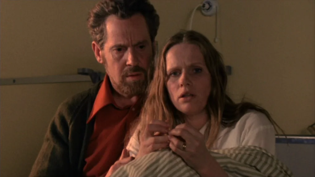 Face to Face (1976) movie still: medium — A hospital room with pale walls and a light switch visible on the wall. A man and a woman sit closely…