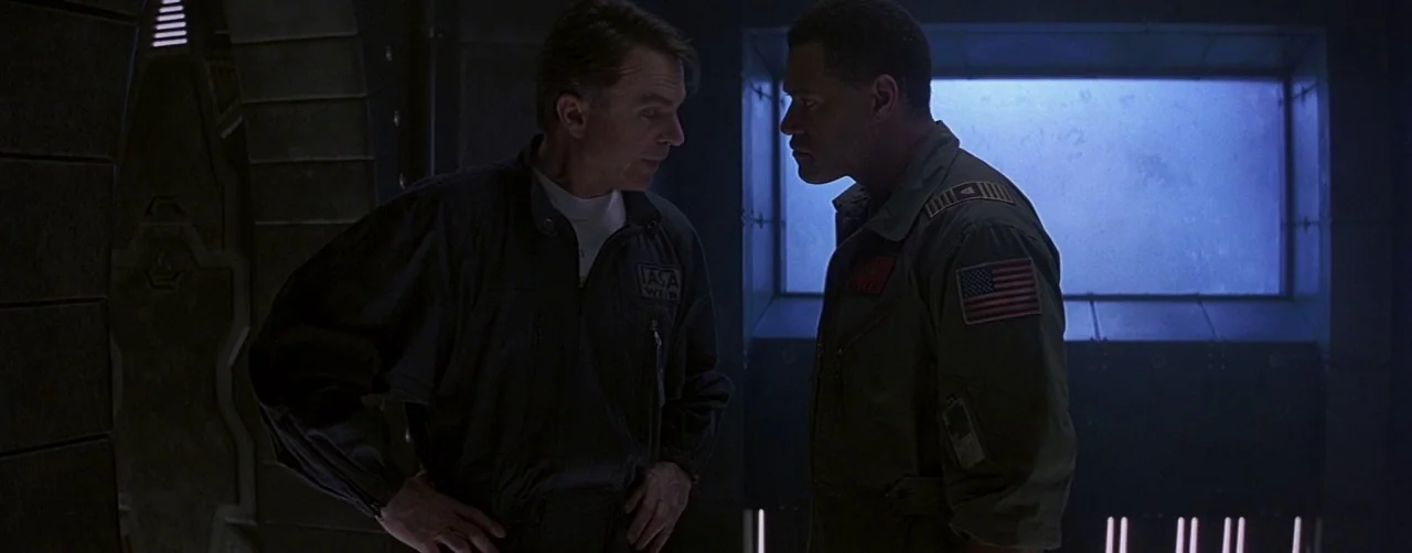 Event Horizon (1997) movie still: medium — A dimly lit industrial space with textured dark walls and a glowing blue viewport. Two men in flight…