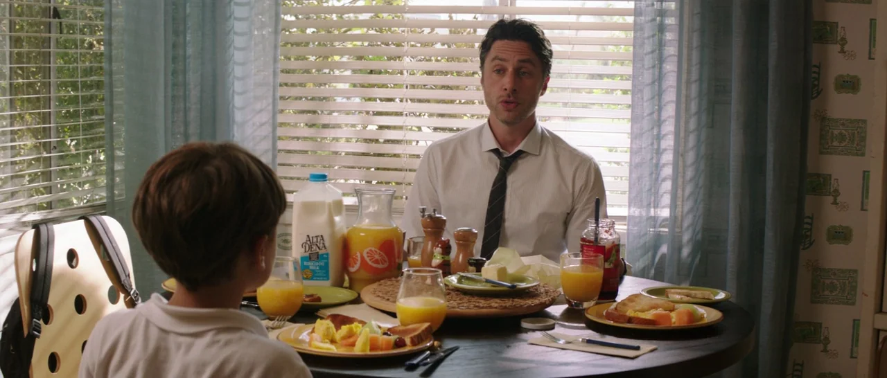 Wish I Was Here (2014) movie still: medium — A sunlit dining room features a round table set for breakfast with plates of food. A man in a dress…