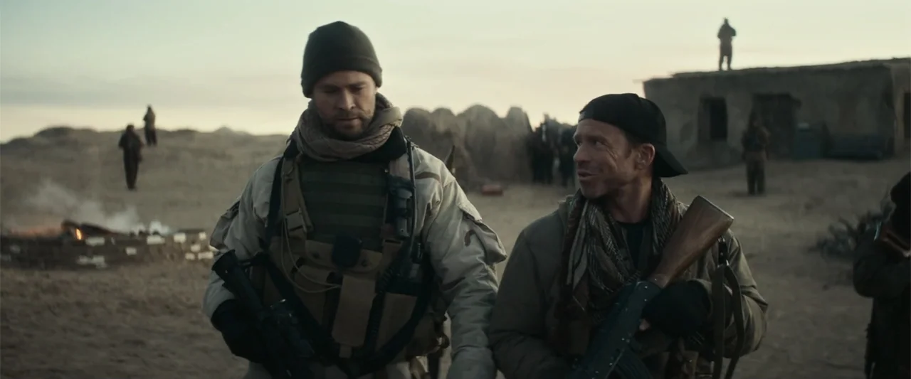 12 Strong (2018) movie still: medium — A dusty desert landscape featuring sparse vegetation and distant mud-brick structures under a sunset. Two…