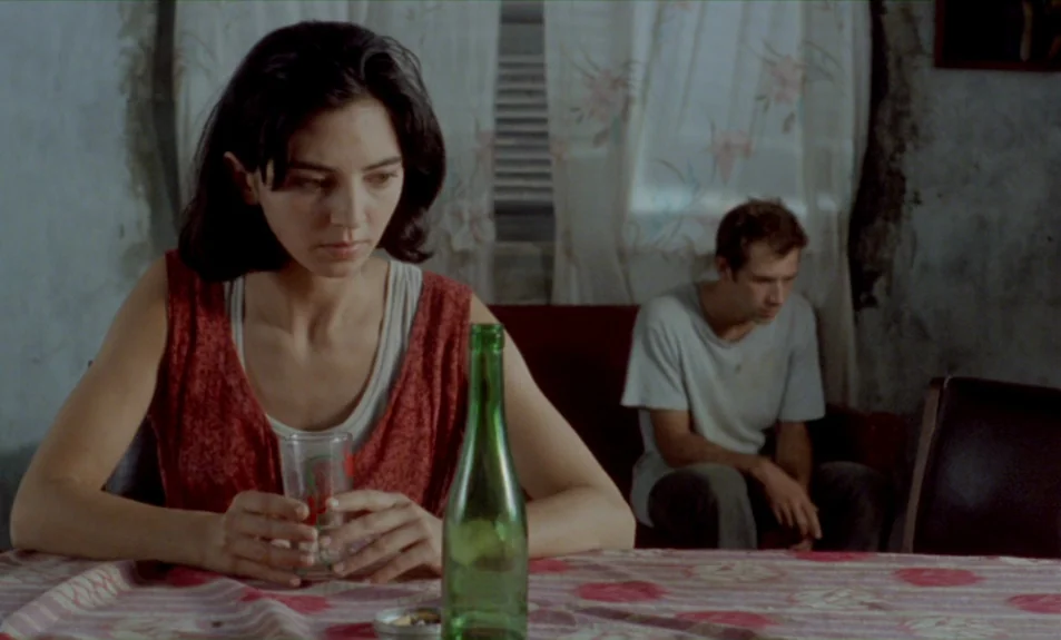 Casa De Lava (1994) movie still: medium — A worn room features a floral patterned tablecloth and white curtains over a window. A woman sits in the…