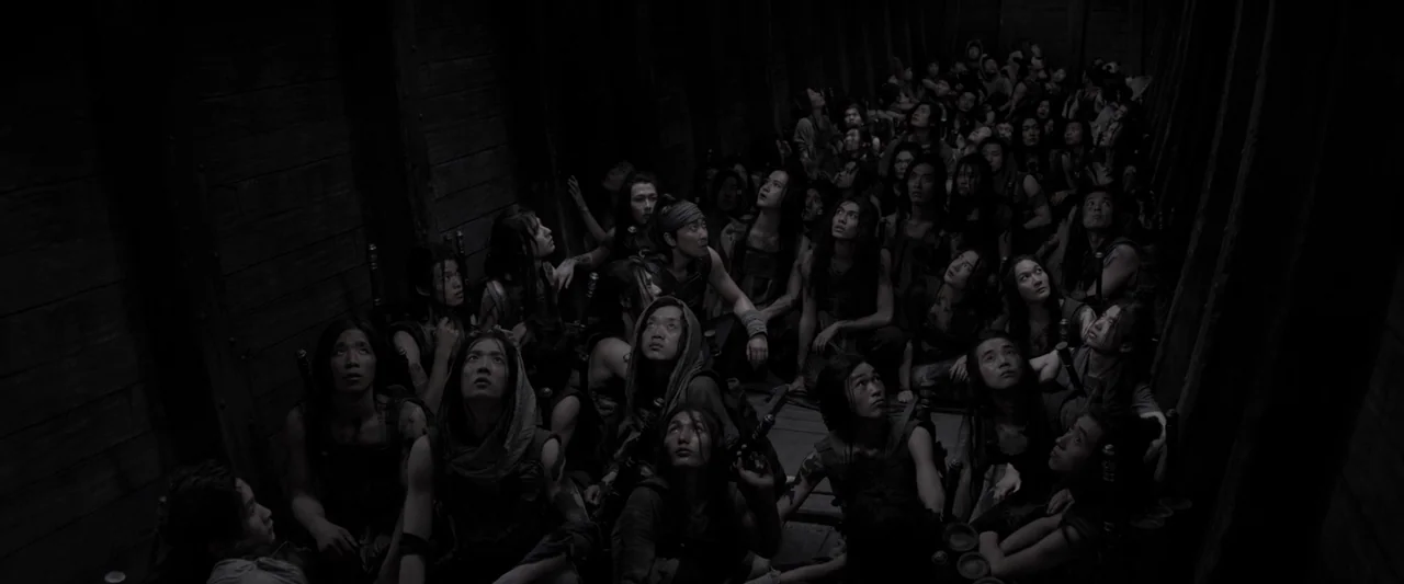 Shadow (2018) movie still: wide — A dark wooden hold of a ship contains a large crowd of people. They sit packed together on the floor while looking…